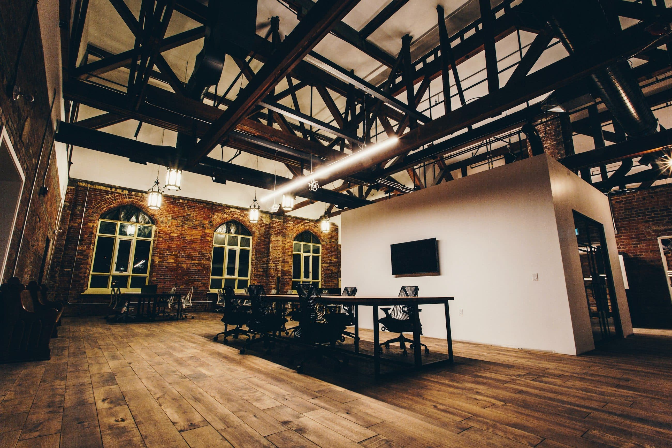 Top-floor office in a former brick warehouse, with exposed timber roof trusses, arched windows and wide plank floors.