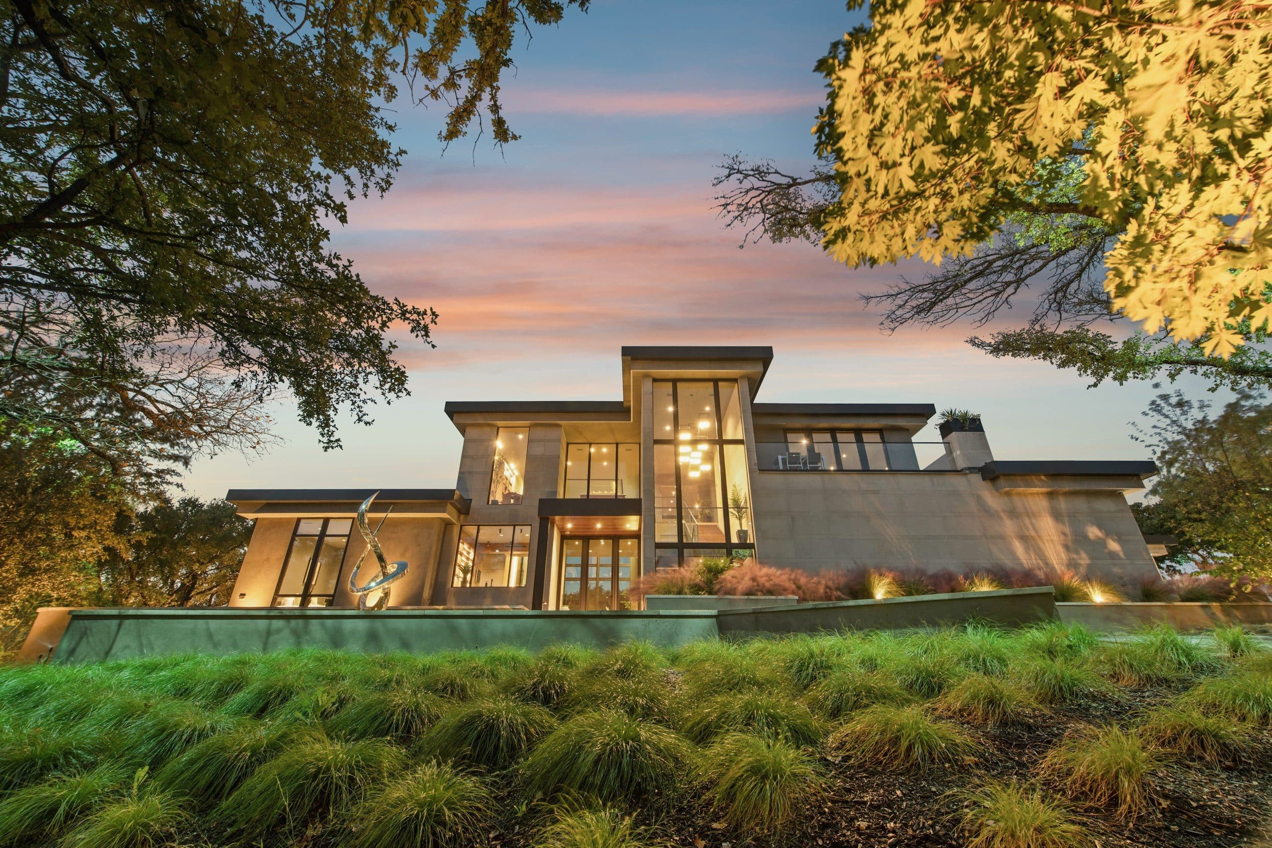 A contemporary stone and glass home at dusk, lit from within, seen across ornamental grasses and framed by oak trees.