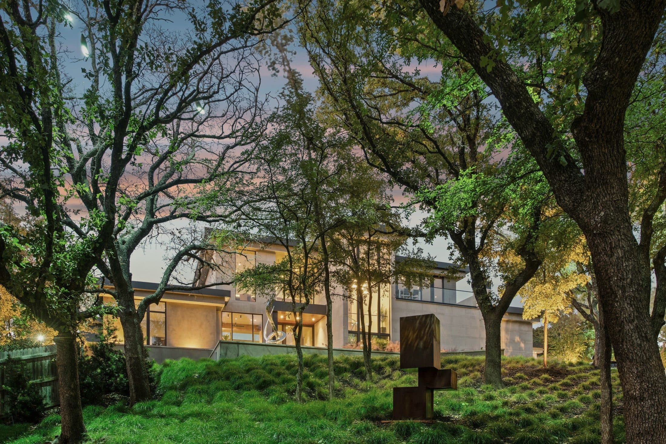 The same house seen through mature oak trees on a sloping lot, with warm light in the tall windows.