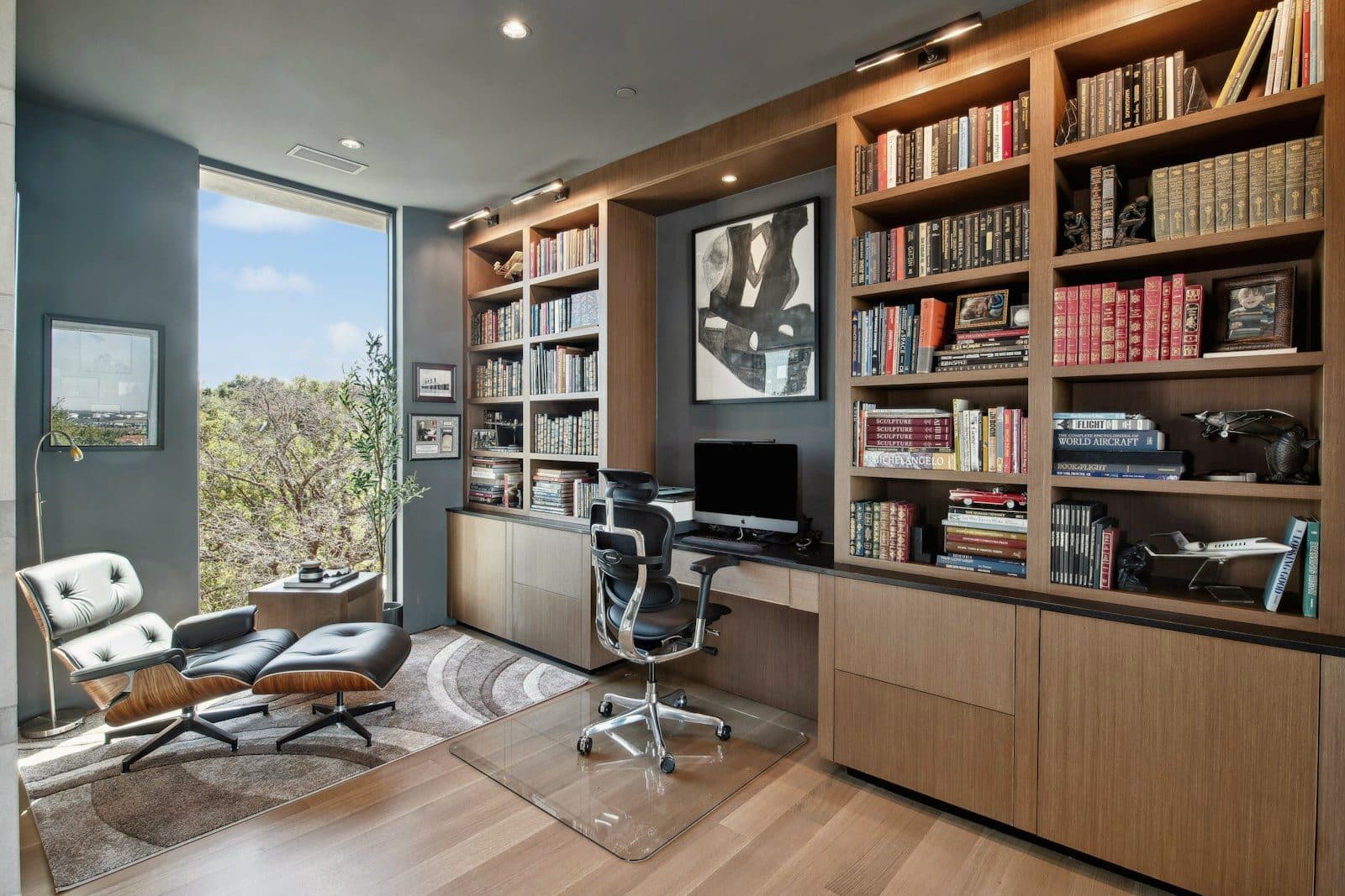 A study with floor-to-ceiling oak shelving and cabinetry beside a tall window looking into the trees.