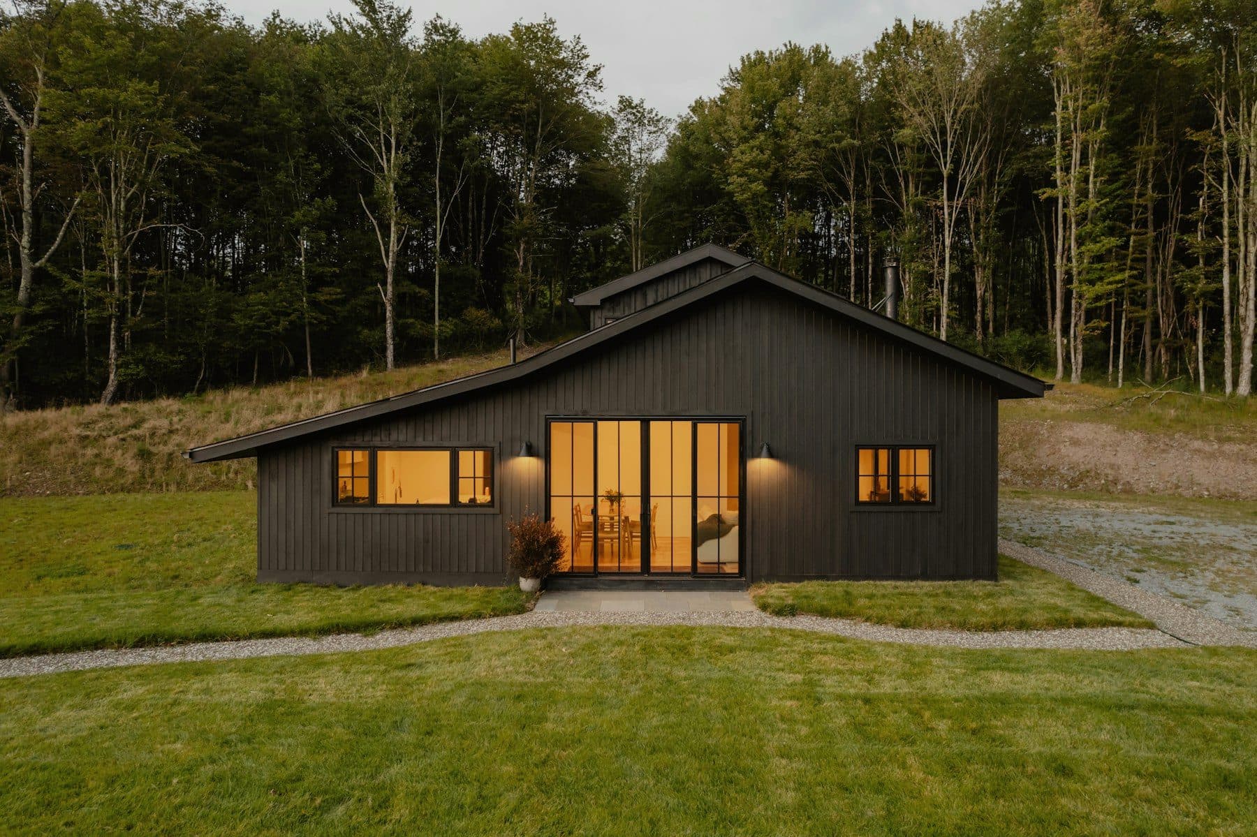 A dark-clad new house with a low gabled roof and tall glazed doors, lit from inside at dusk at the edge of a wood.